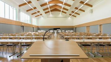 Un amplio salón de conferencias con filas de mesas y sillas, un atril, y un techo con vigas de madera e iluminación.