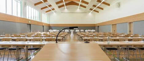 Un amplio salón de conferencias con filas de mesas y sillas, un atril, y un techo con vigas de madera e iluminación.