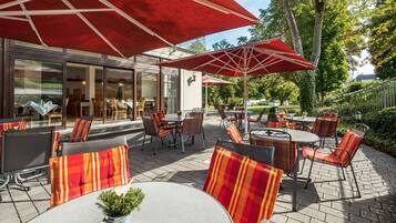 Ein Außenbereich mit Terrasse, Tischen und Stühlen, beschattet durch rote Sonnenschirme, mit Blick ins Gebäudeinnere.