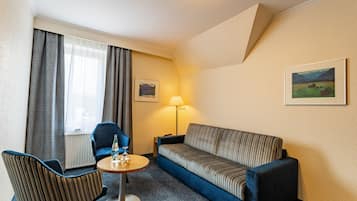Een compacte hotelkamer met een blauwe bank, twee fauteuils, een klein rond tafeltje, een lamp en twee ingelijste foto's aan de muur.