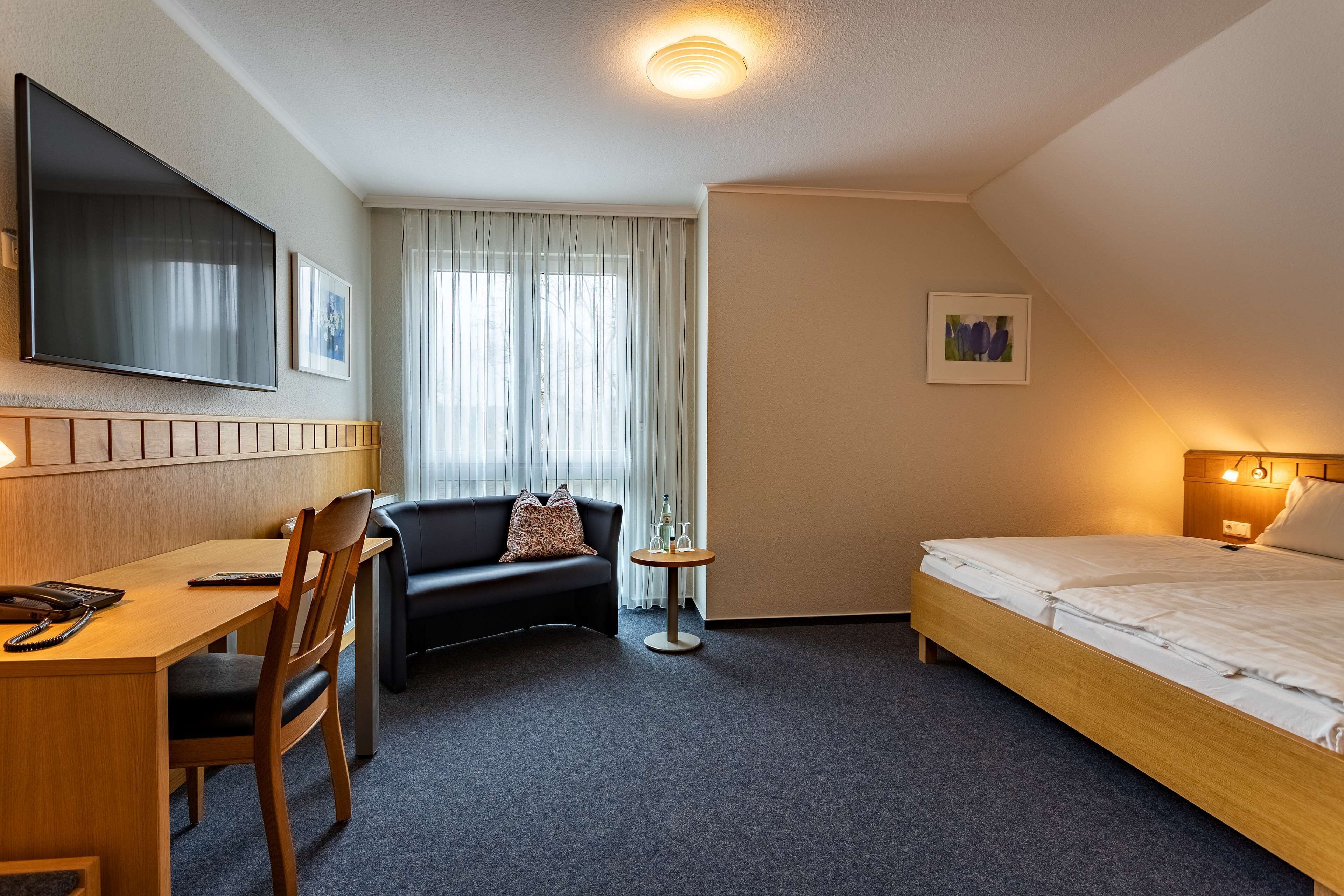 A hotel room with a bed, a desk, a chair, a sofa, a TV, and a window with curtains.