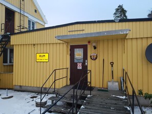 A yellow building with a wooden staircase leading to a door, a sign reading "RUM 070 88 03560", and a no-smoking sign.