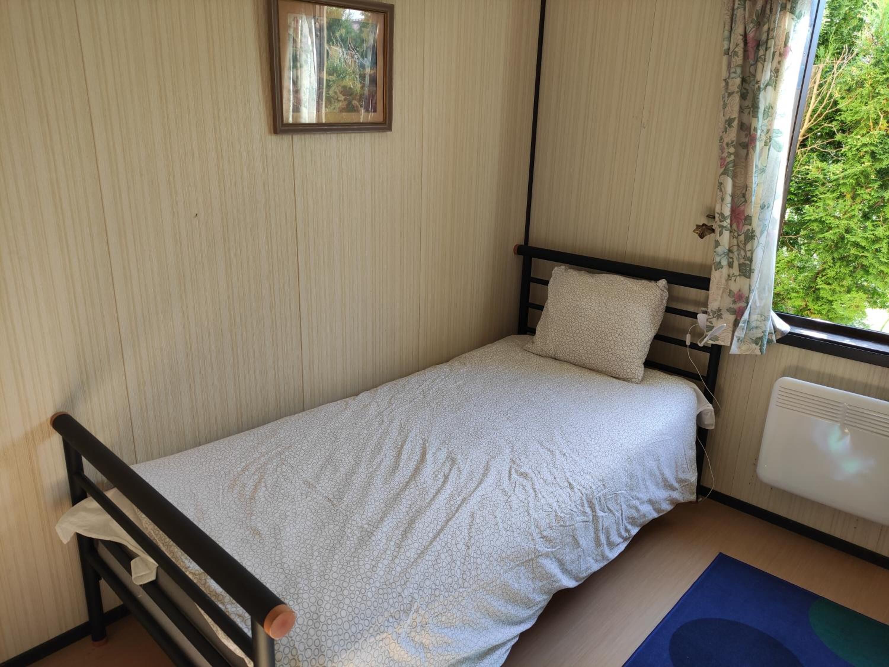 Una habitación pequeña con cama individual, estructura de madera, ventana con cortinas florales y un radiador.