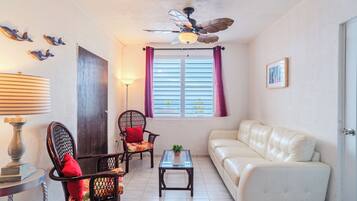 A living room with a white sofa, two wooden chairs, a glass coffee table, a ceiling fan, and a window with curtains.