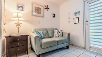 Una habitación con un sofá color beige, una pequeña mesita de noche con cajones, una lámpara en forma de estrella y una puerta con persianas horizontales.