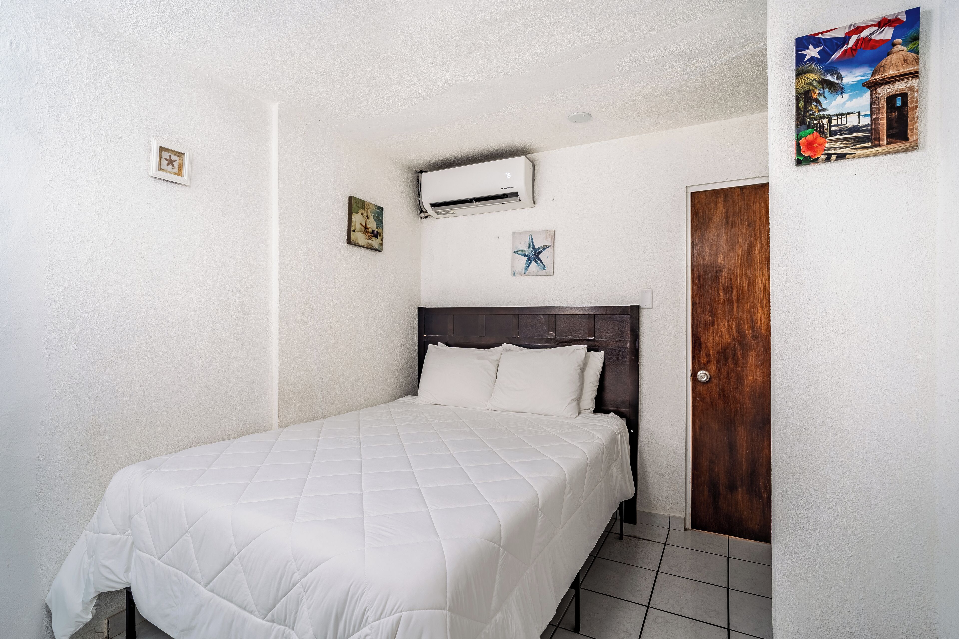A bedroom with a bed, a wooden door, an air conditioner, and a wall-mounted picture.