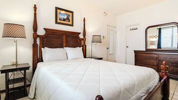 A neatly made bed with a wooden headboard, a bedside table with a lamp, a dresser with drawers, and a framed picture on the wall.