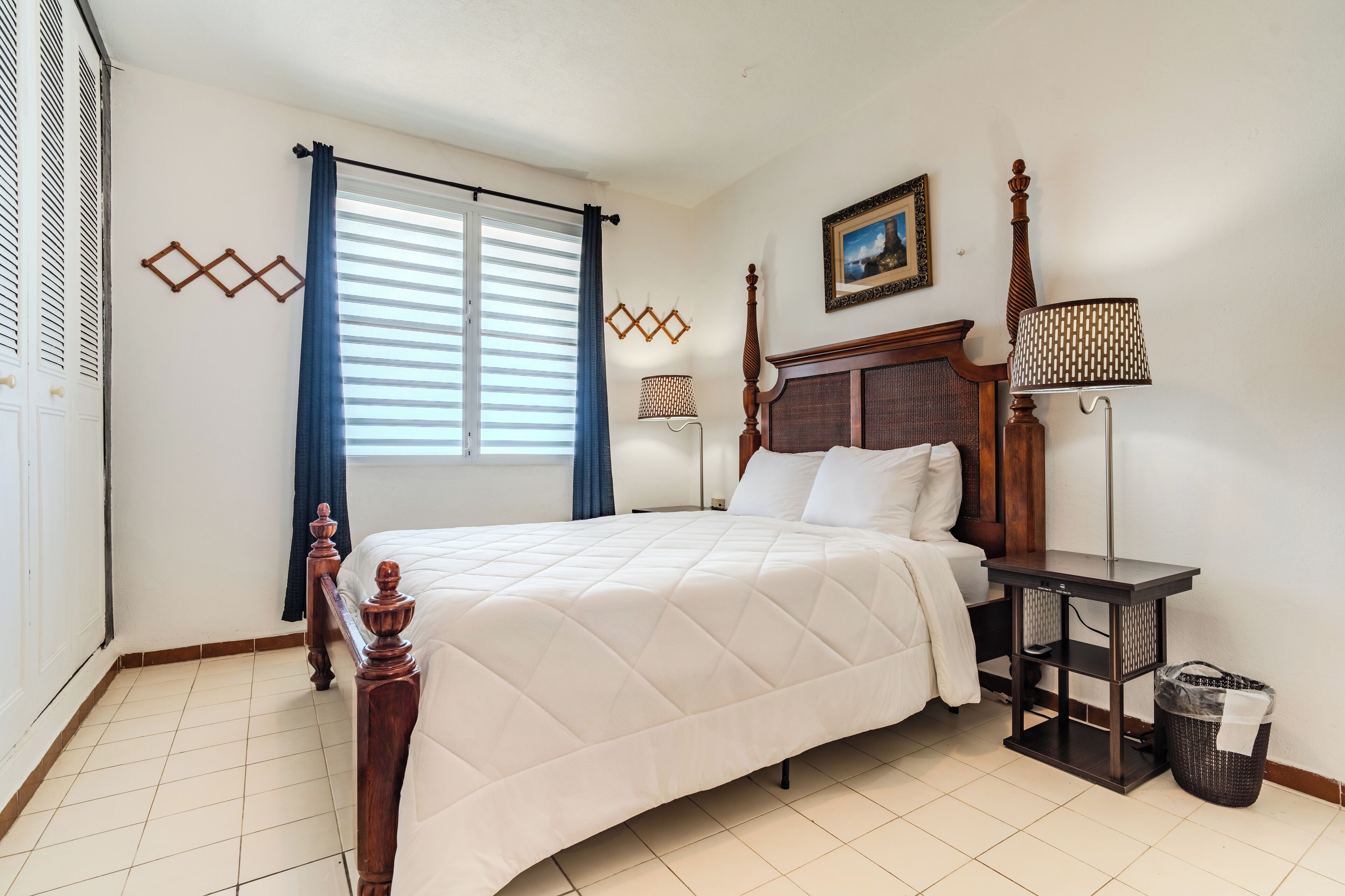 A neatly arranged bedroom with a bed, a bedside table with a lamp, a picture on the wall, and a window with curtains.