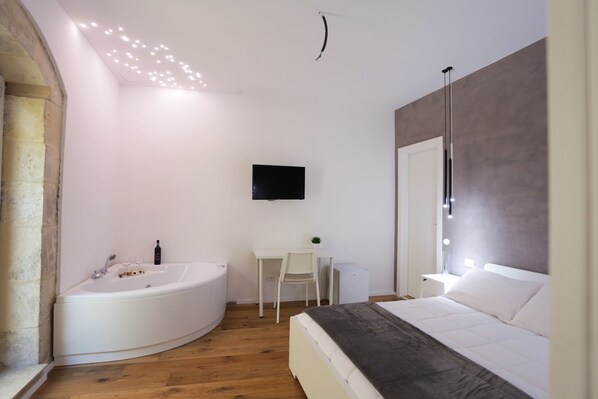 A modern bedroom with a large bed, a built-in bathtub, a desk, and a TV mounted on the wall. - Reusuite Authentic Sicilian Hospitality (Ragusa)