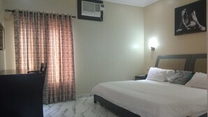 Quarto de hotel com cama, escrivaninha, cadeira, cortina, aparelho de ar condicionado e um quadro na parede.