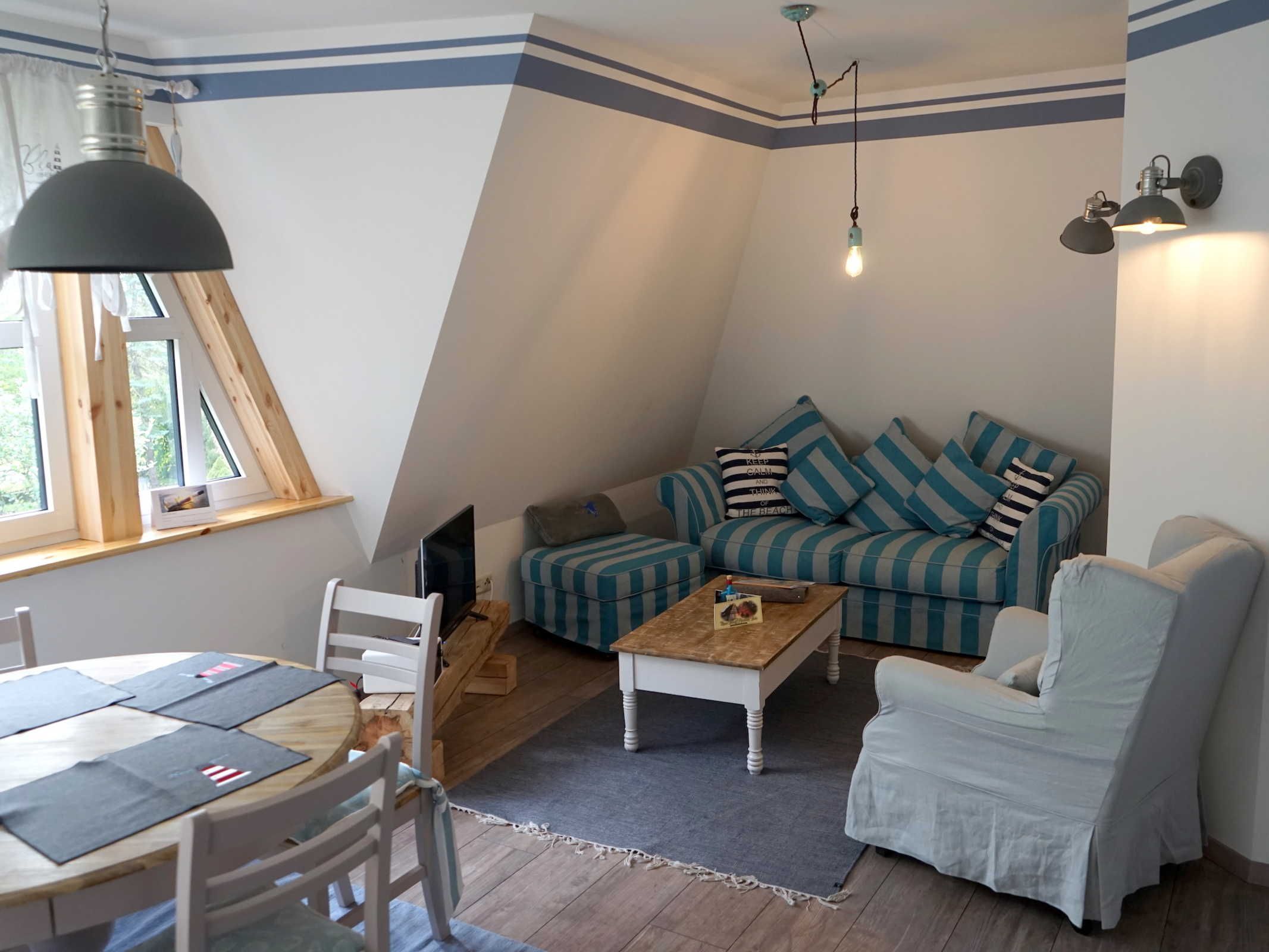 A cozy loft-style living room with a sloped ceiling, a striped sofa, a white armchair, a wooden coffee table, and a dining table with chairs.