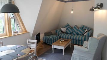 Ein gemütliches Loft-Wohnzimmer mit schrägem Dach, einem gestreiften Sofa, einem weißen Sessel, einem Holztisch und einem Esstisch mit Stühlen.