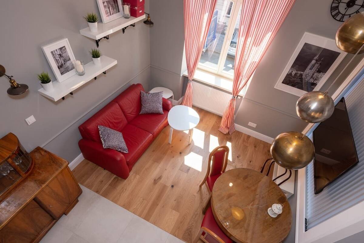 A modern living room with a red sofa, a round wooden dining table, and a small white table. There are shelves with decorative items and a window with curtains.