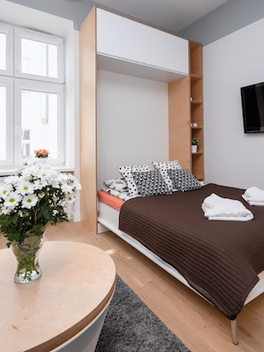 Ein modernes Schlafzimmer mit Bett, einem kleinen Tisch mit Blumenvase, einem an der Wand montierten Fernseher und einem Fenster mit Blick nach draußen.