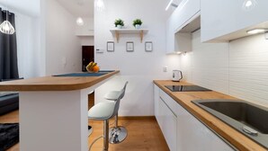 Une cuisine moderne avec des armoires blanches, un comptoir en bois et un évier encastré. On y trouve deux tabourets de bar et un petit îlot central sur lequel est posé un panier de fruits.