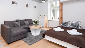 Une pièce moderne et bien éclairée, avec un canapé gris, une table basse ronde, un lit agrémenté d’oreillers à motifs et une fenêtre donnant sur des arbres.