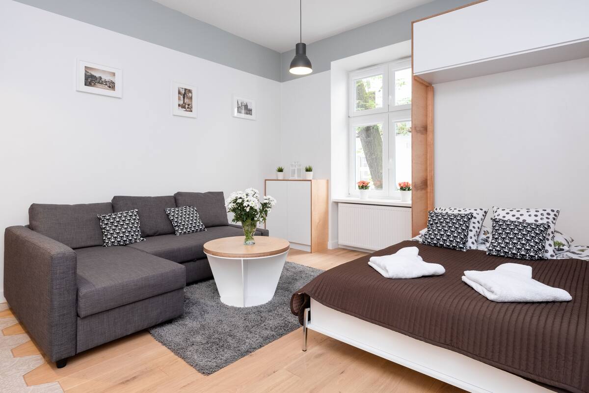 A modern, well-lit room with a grey sofa, a round coffee table, a bed with patterned pillows, and a window with a view of trees.