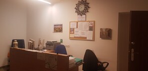 Ein Empfangstresen mit Computer, Drucker und verschiedenen Unterlagen. An der Wand befindet sich eine Uhr und ein Schwarzes Brett mit Aushängen.