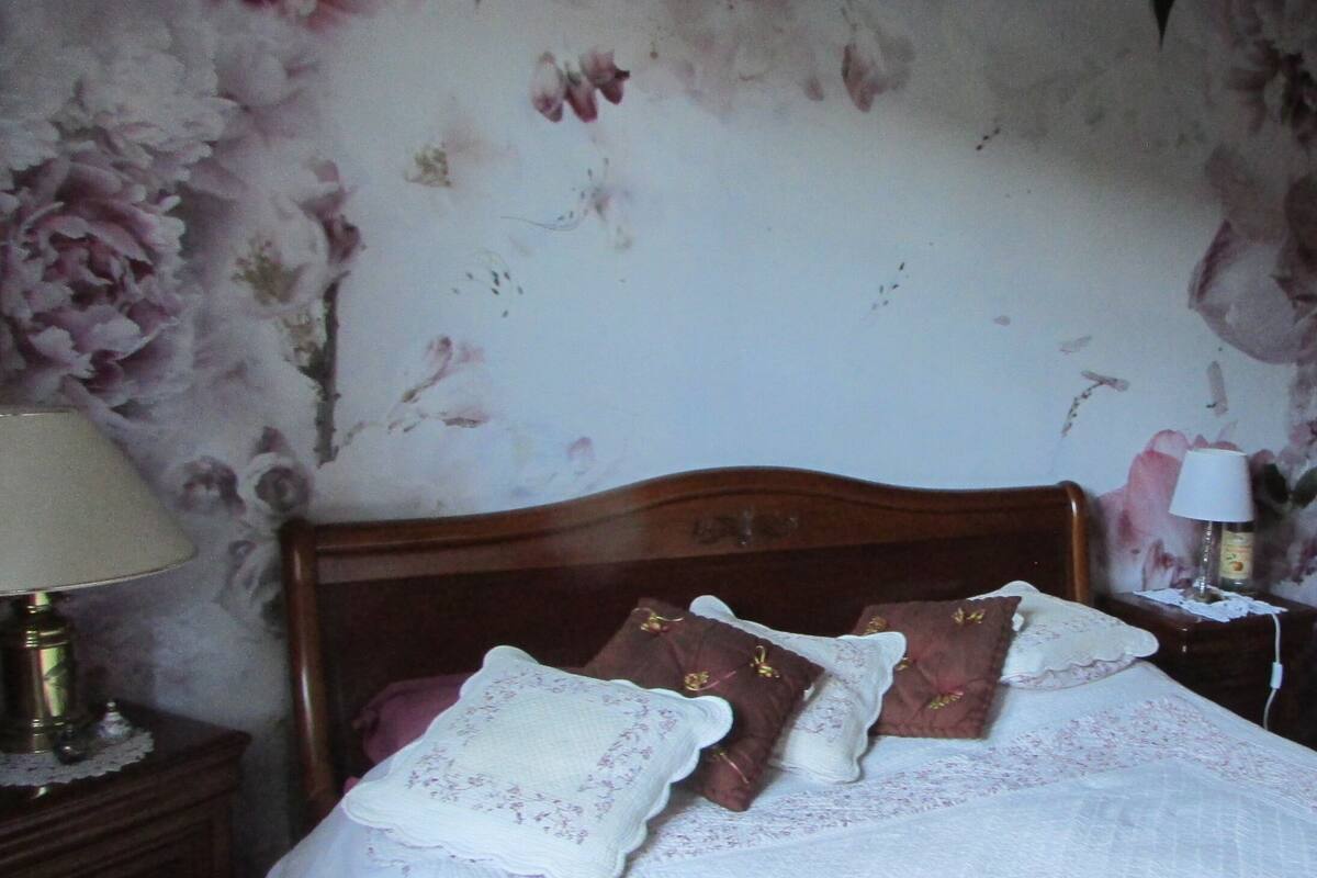A bedroom with a wooden headboard, a bed with white and brown pillows, a nightstand with a lamp, and a floral wallpaper.