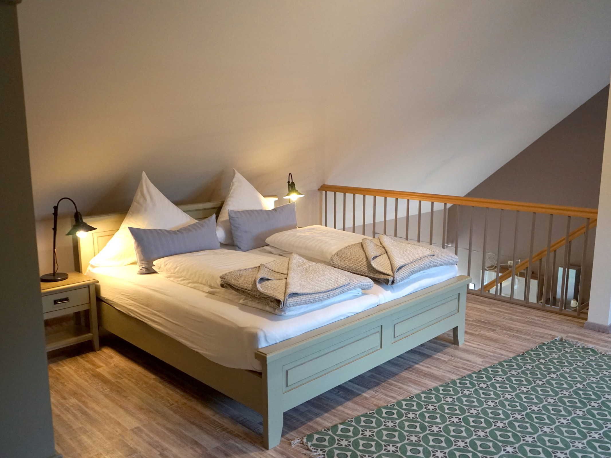 Ein Schlafzimmer mit einem Bett, Kissen, einem Nachttisch, einer Lampe und einem gemusterten Teppich.