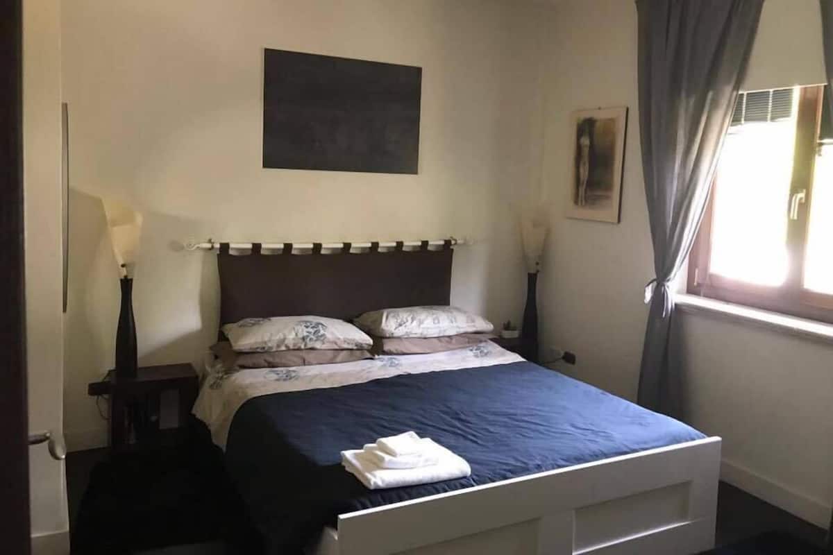 Une chambre à coucher avec un lit, une table de chevet, une lampe, une fenêtre avec des rideaux et un tableau au mur.