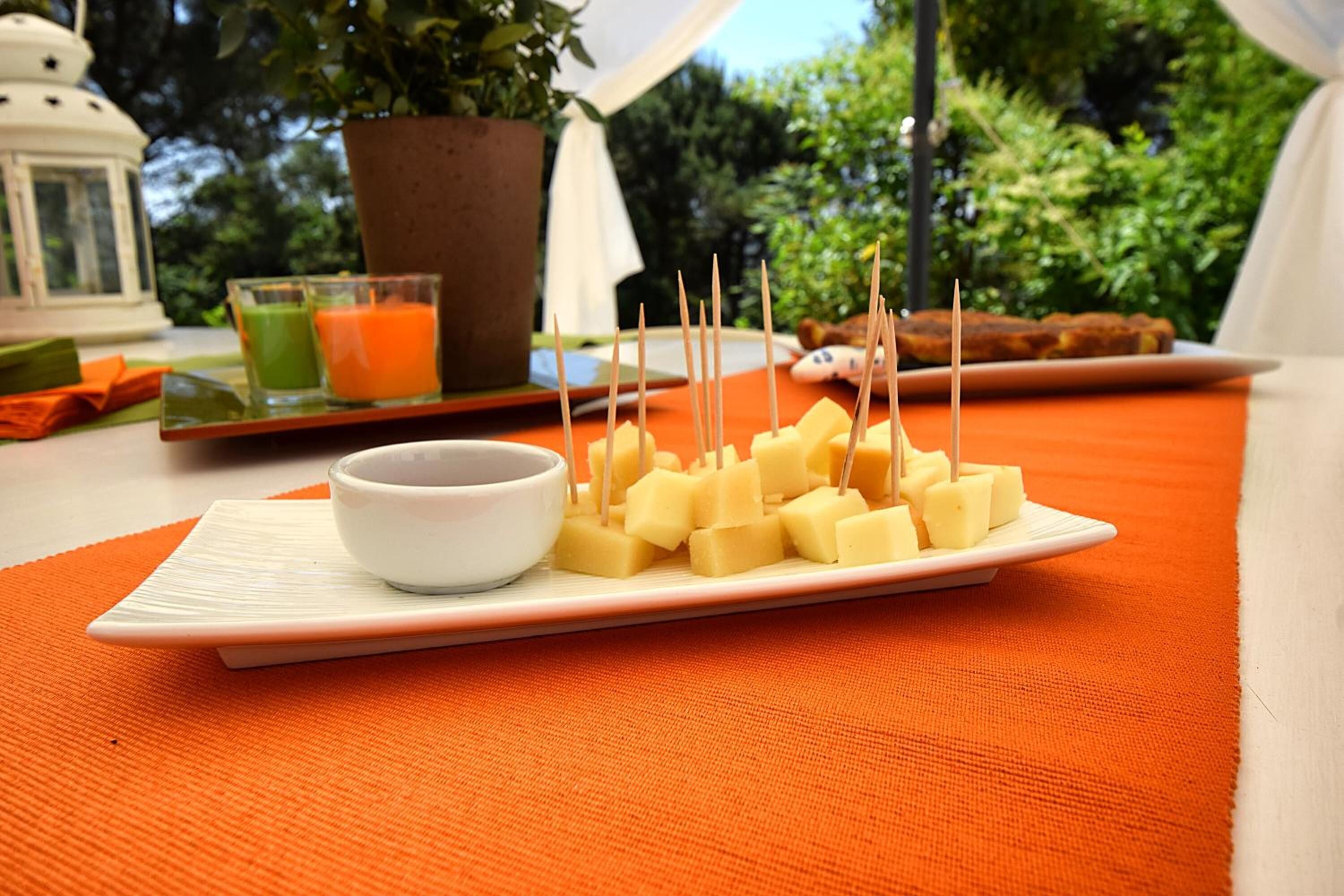 A plate of cheese cubes with toothpicks on an orange placemat, a small white bowl, and two glasses of juice on a table with a potted plant and a lantern in the background.