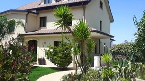 Une maison à deux étages, avec un toit recouvert de tuiles, une antenne parabolique et un jardin bien entretenu, agrémenté de diverses plantes et d’un chemin menant à l’entrée.