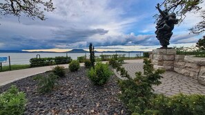 A landscaped area with a statue, a paved walkway, and a view of a body of water and distant mountains.