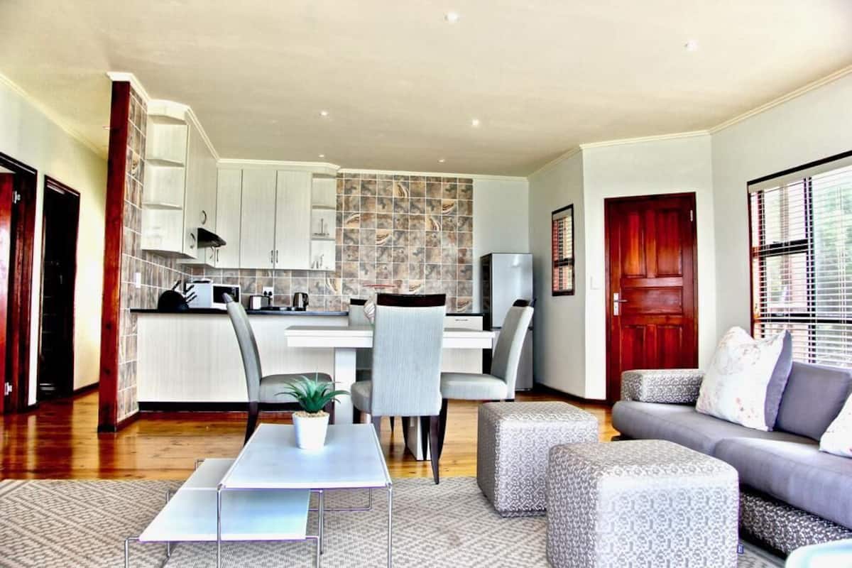 A modern living room with a grey sofa, a dining table with chairs, and a kitchen area with white cabinets and a tiled backsplash.