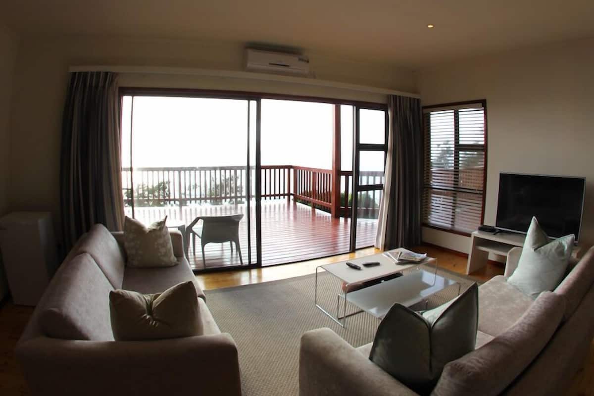 A spacious living room with a large sliding glass door leading to an outdoor balcony. There is a sofa set, a coffee table, and a flat-screen TV.
