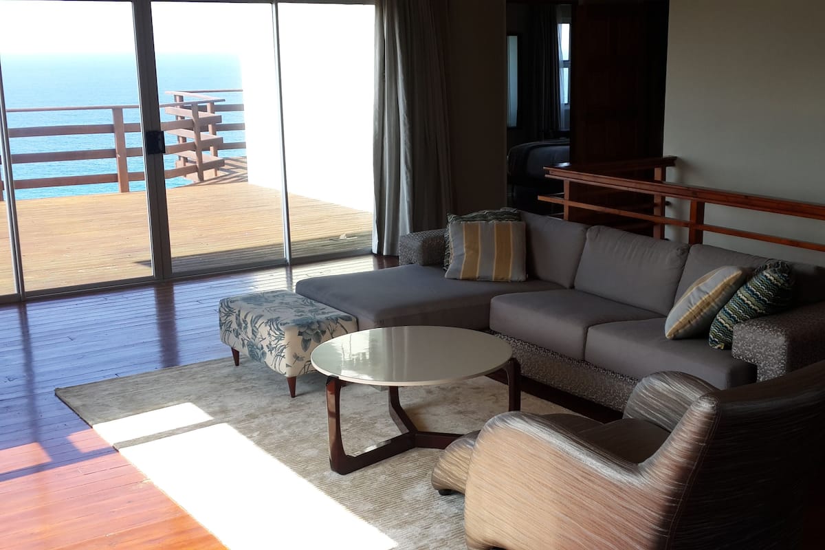 A modern living room with a large sofa, a small round table, and a patterned ottoman. A wooden deck with a railing is visible through the glass doors.