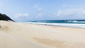 Una amplia playa de arena con olas suaves, cielo despejado y una vista distante del océano.
