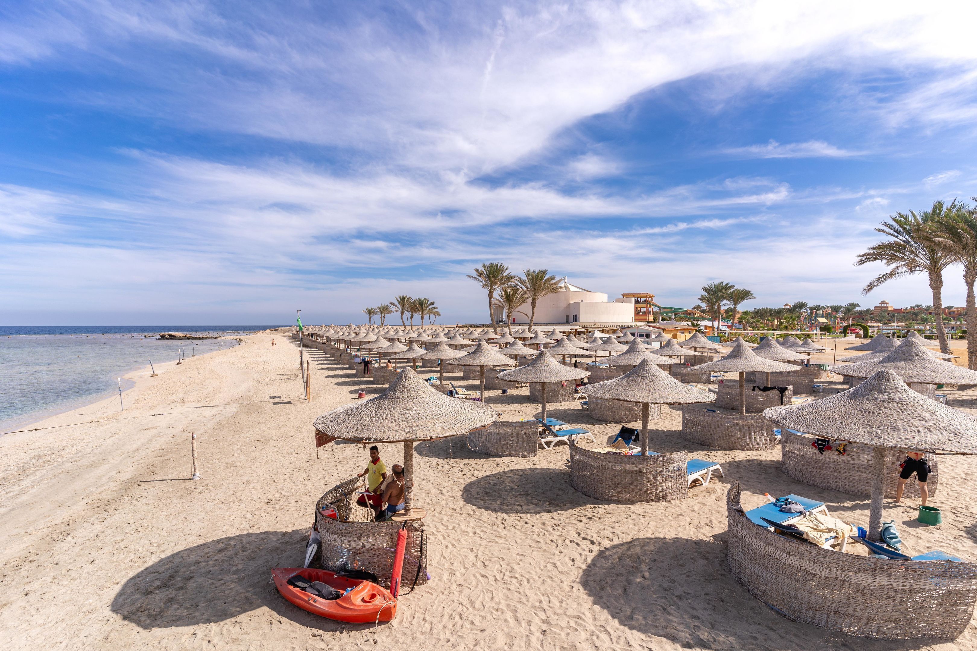 A beach with thatched umbrellas and lounge chairs, a sandy shore, and a building in the background.