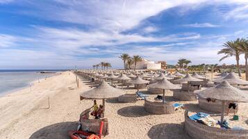 A beach with thatched umbrellas and lounge chairs, a sandy shore, and a building in the background.