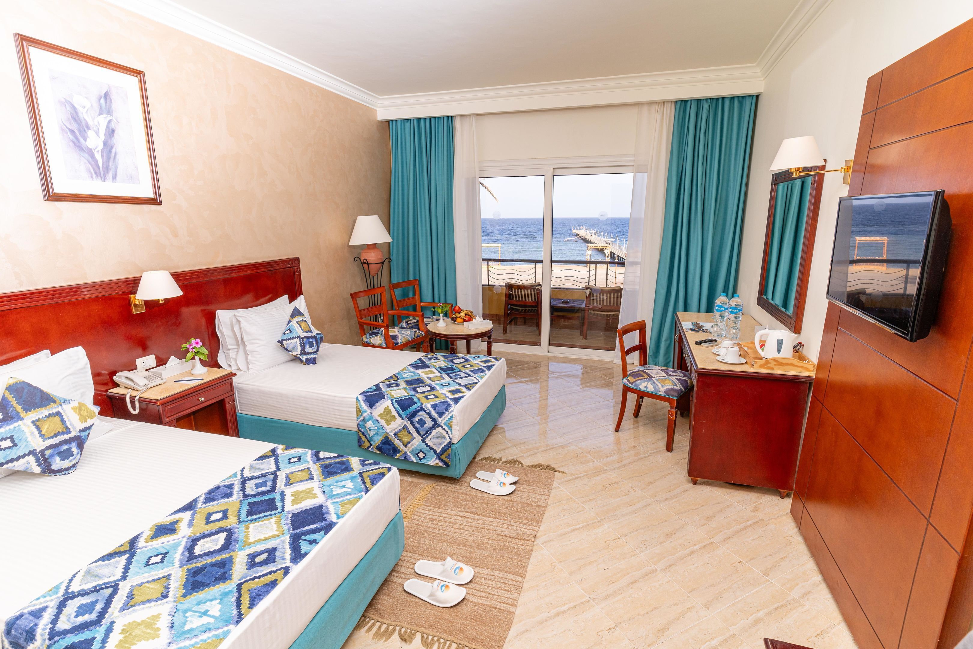Habitación de hotel con dos camas, un escritorio, una silla, un televisor, un balcón con vista al mar y una mesa con sillas.
