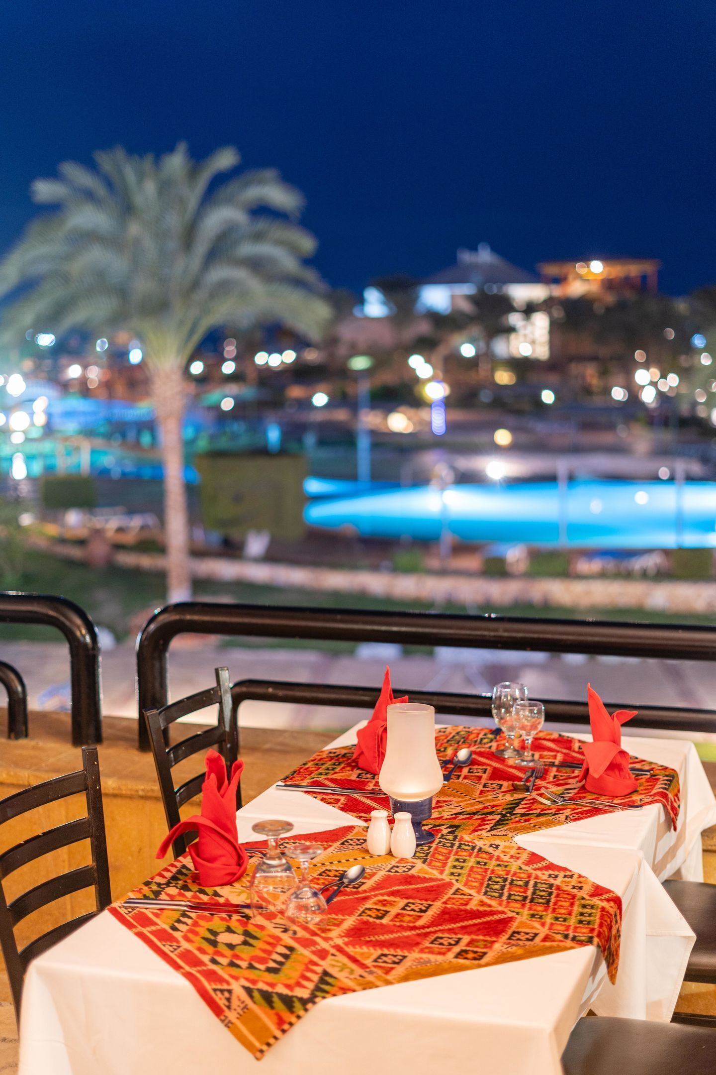 Mesa de comedor dispuesta con mantel estampado, servilletas rojas y cristalería, con vista a una piscina y edificios de noche.