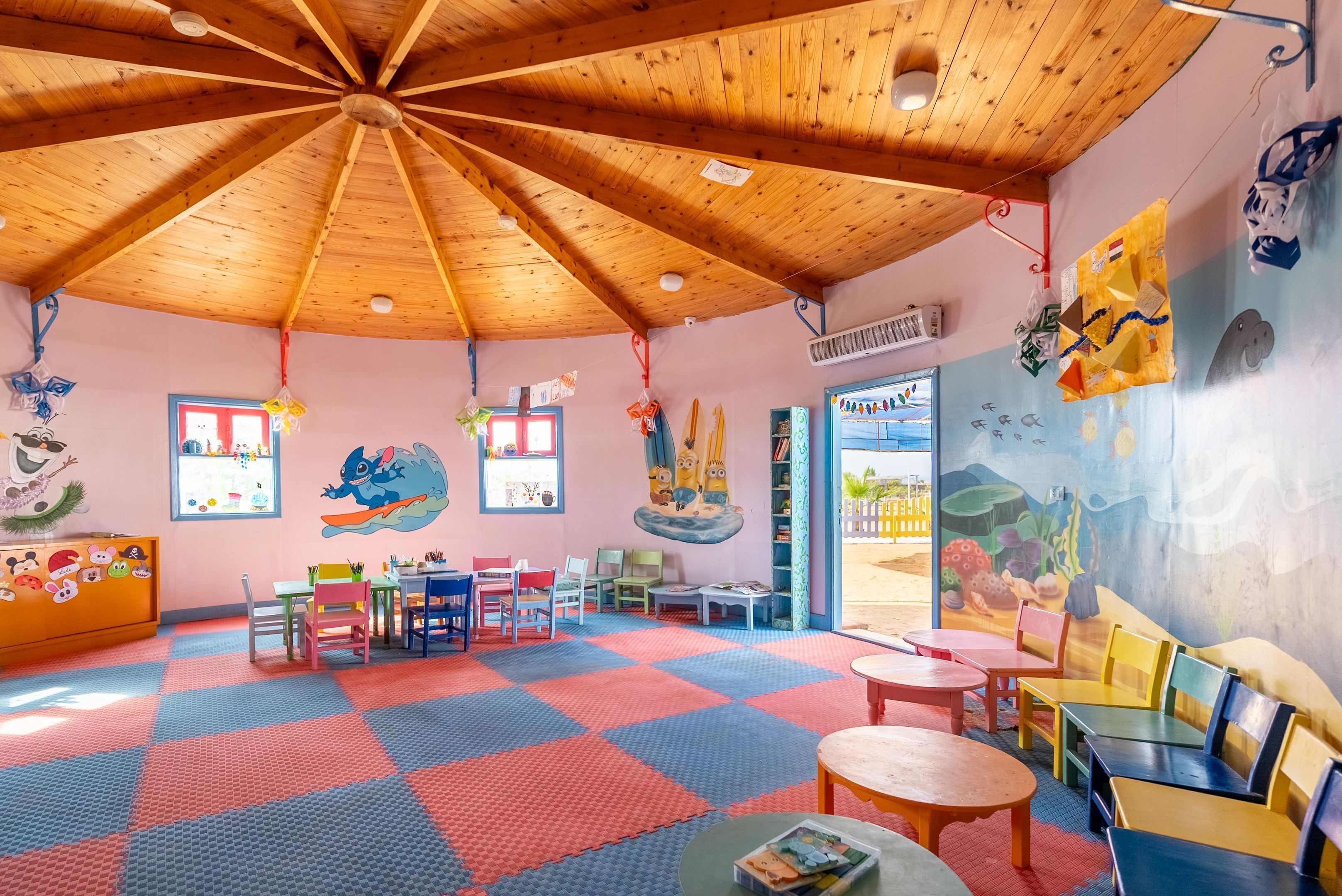 Une salle de jeux pour enfants avec un plafond en bois, un tapis de sol coloré et différentes tables et chaises pour jouer.