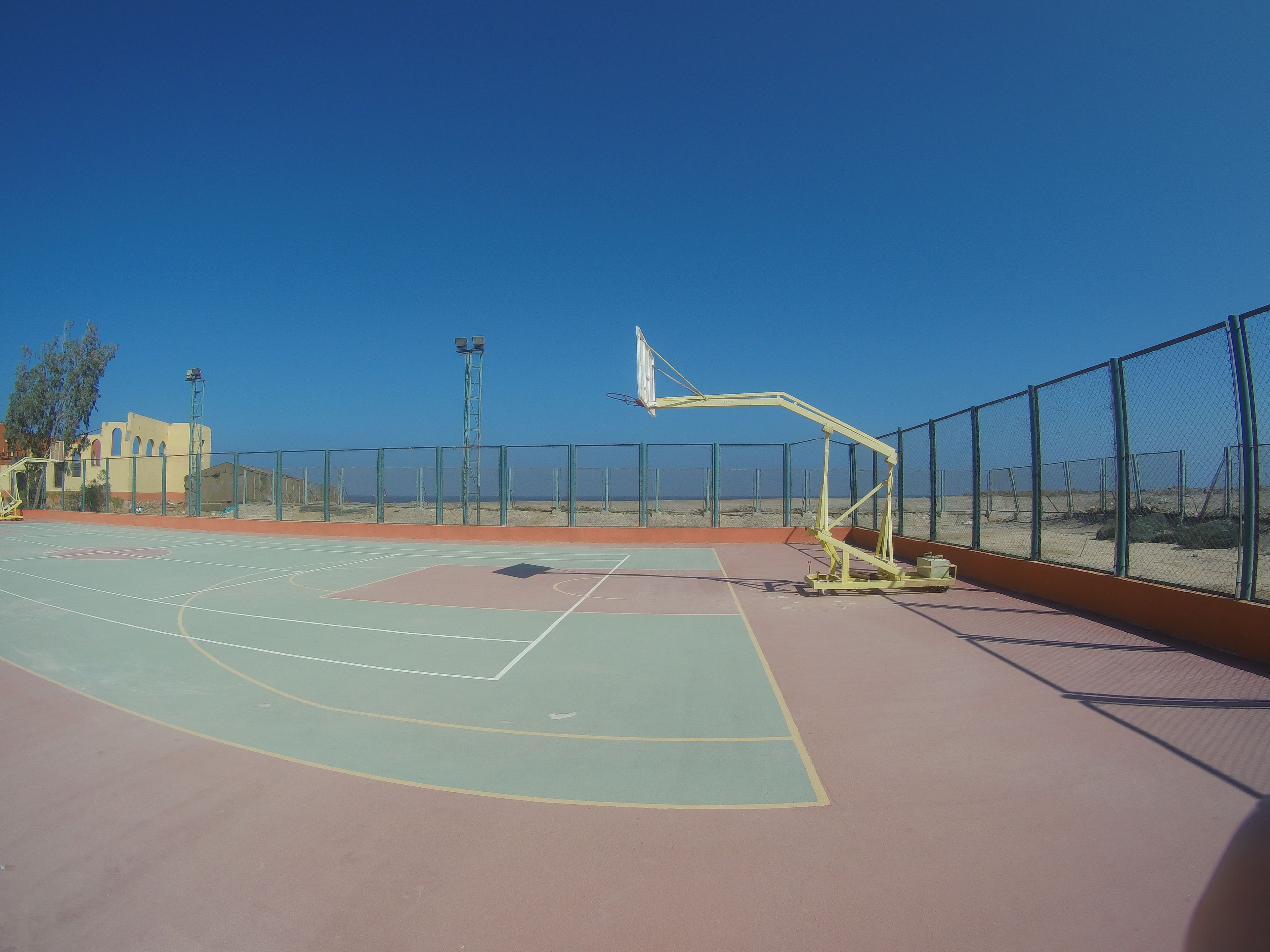 Un terrain de basket en plein air, avec un ciel dégagé, une clôture et un bâtiment en arrière-plan.