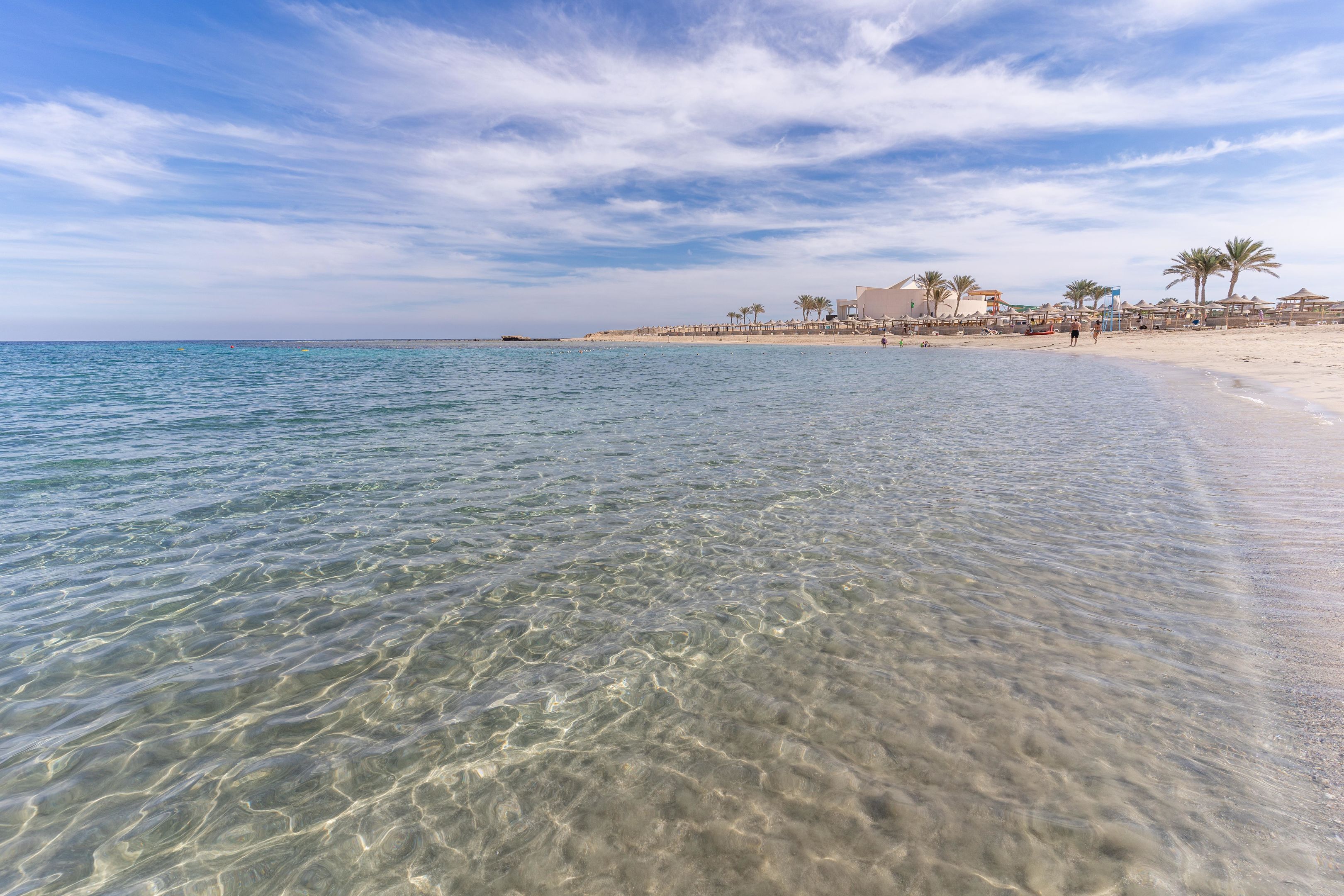 A beach with clear shallow waters, palm trees, and a few buildings in the background.