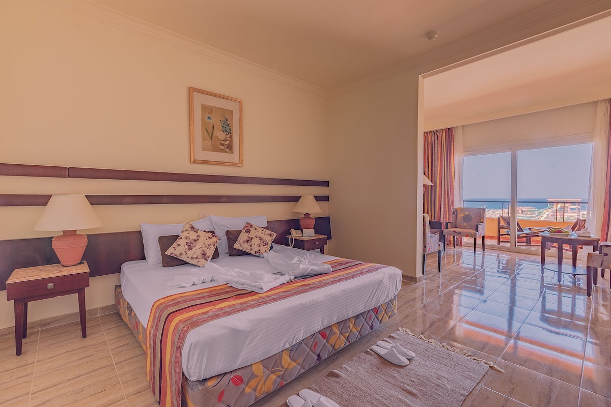A hotel room with a bed, two bedside lamps, a nightstand, a large window with a view, and a balcony area.