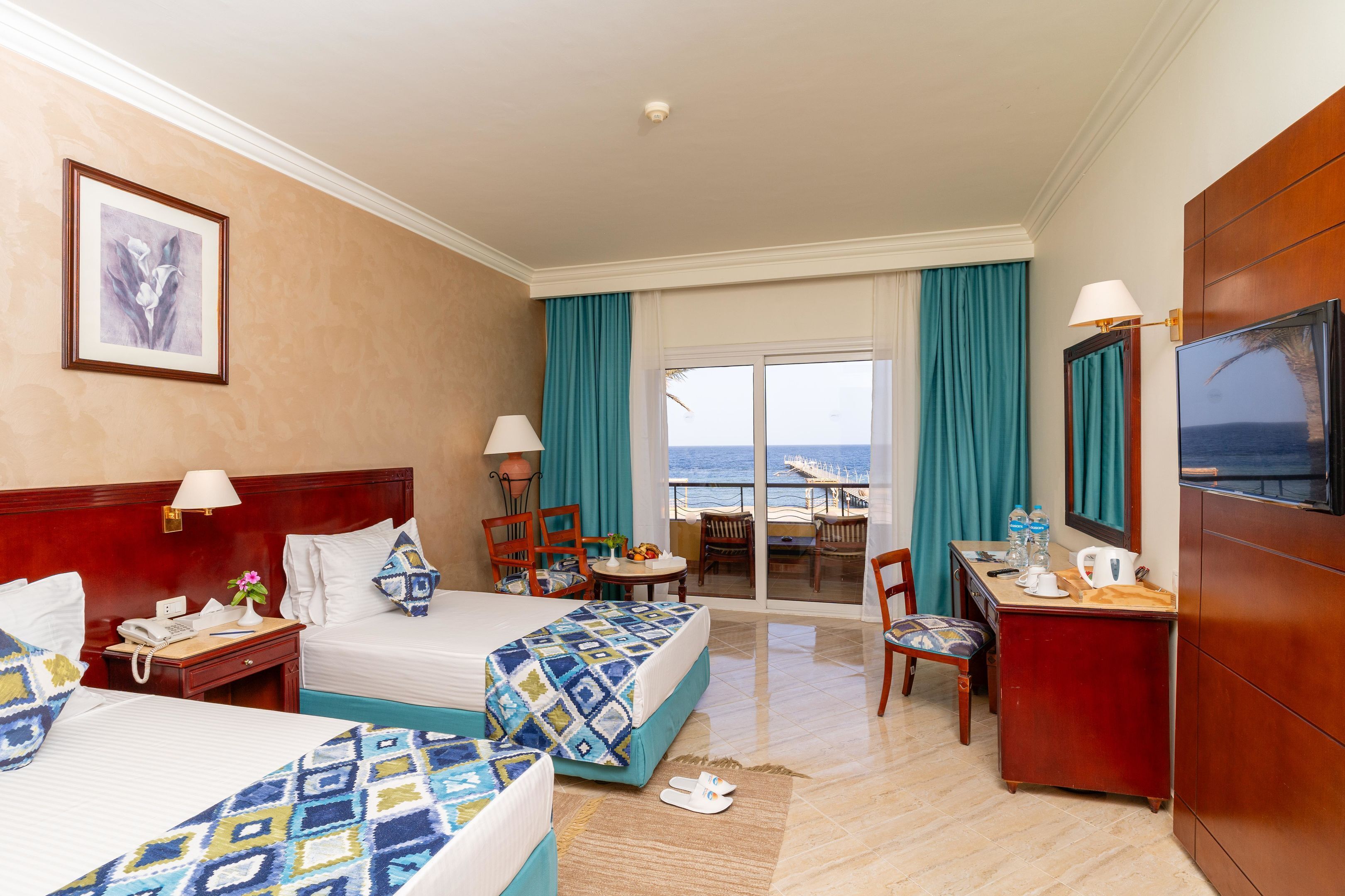 Een hotelkamer met twee bedden, een bureau, een stoel, een televisie en een balkon met uitzicht op zee.