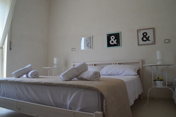 Una cama bien tendida con toallas enrolladas, una mesita de noche con una lámpara y dos cuadros colgados en la pared con texto.