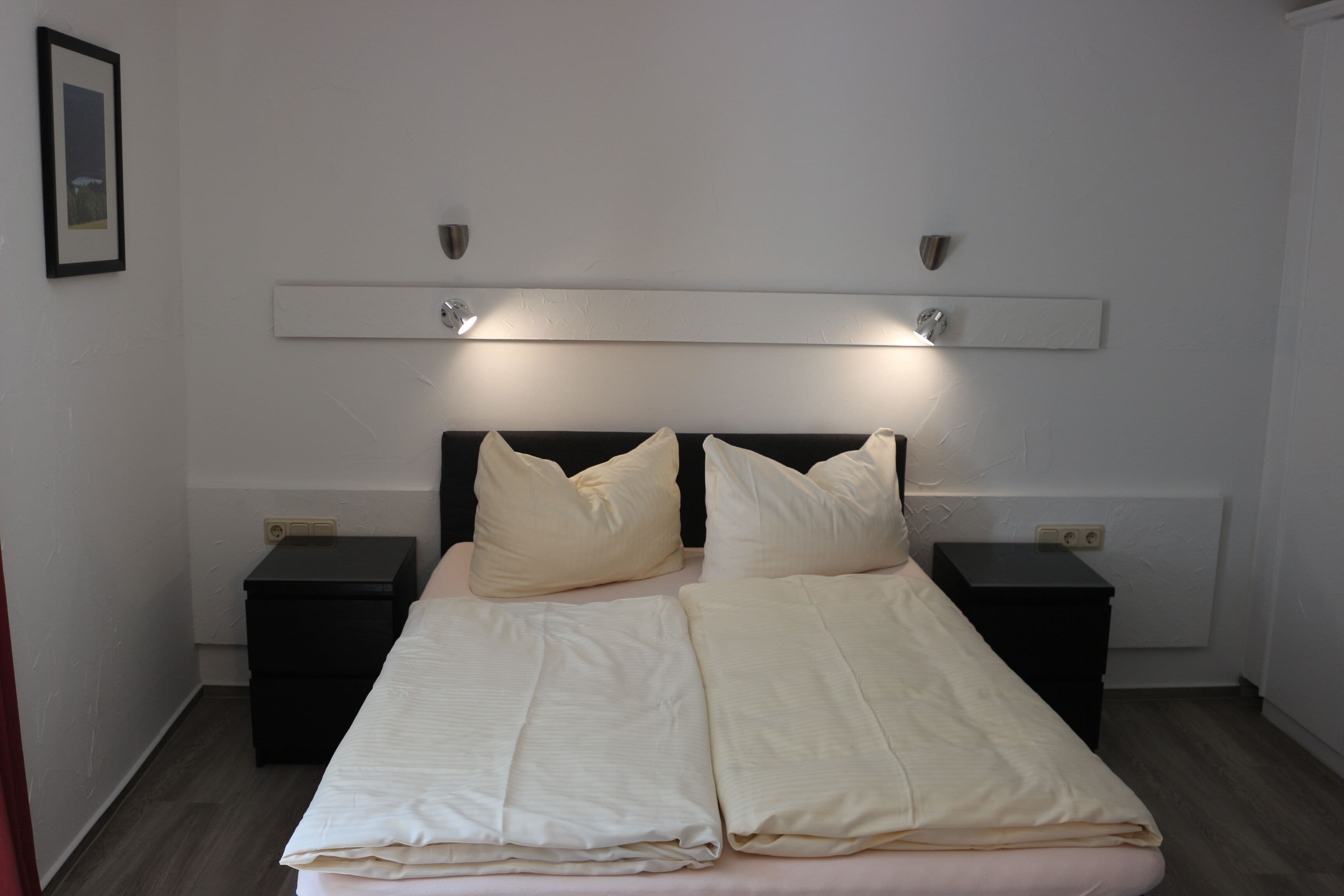 Un lit double avec une couette et des oreillers blancs, deux tables de chevet et un luminaire fixé au mur.
