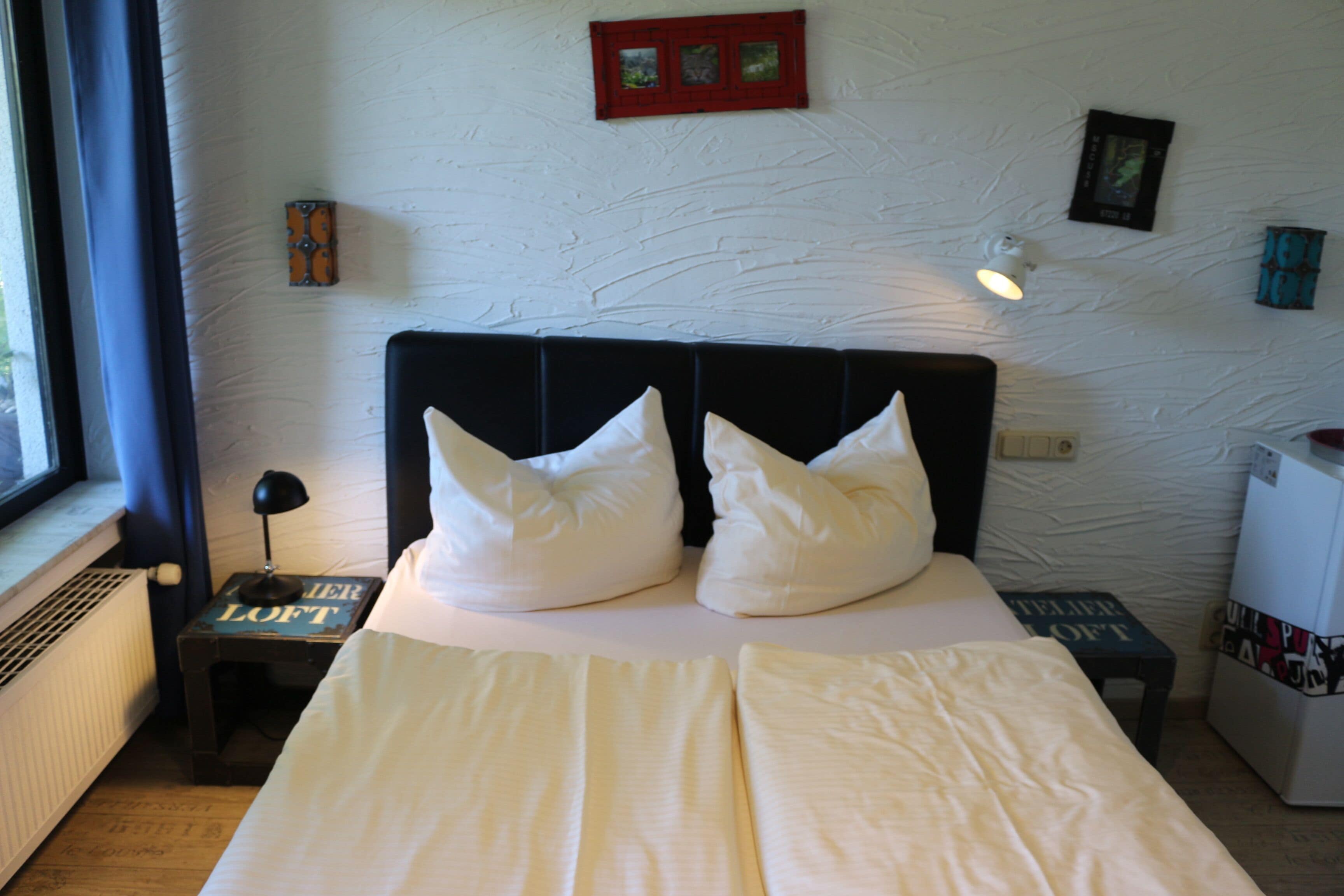 A neatly made bed with white bedding and pillows, a bedside table with a lamp, and a small shelf with books.