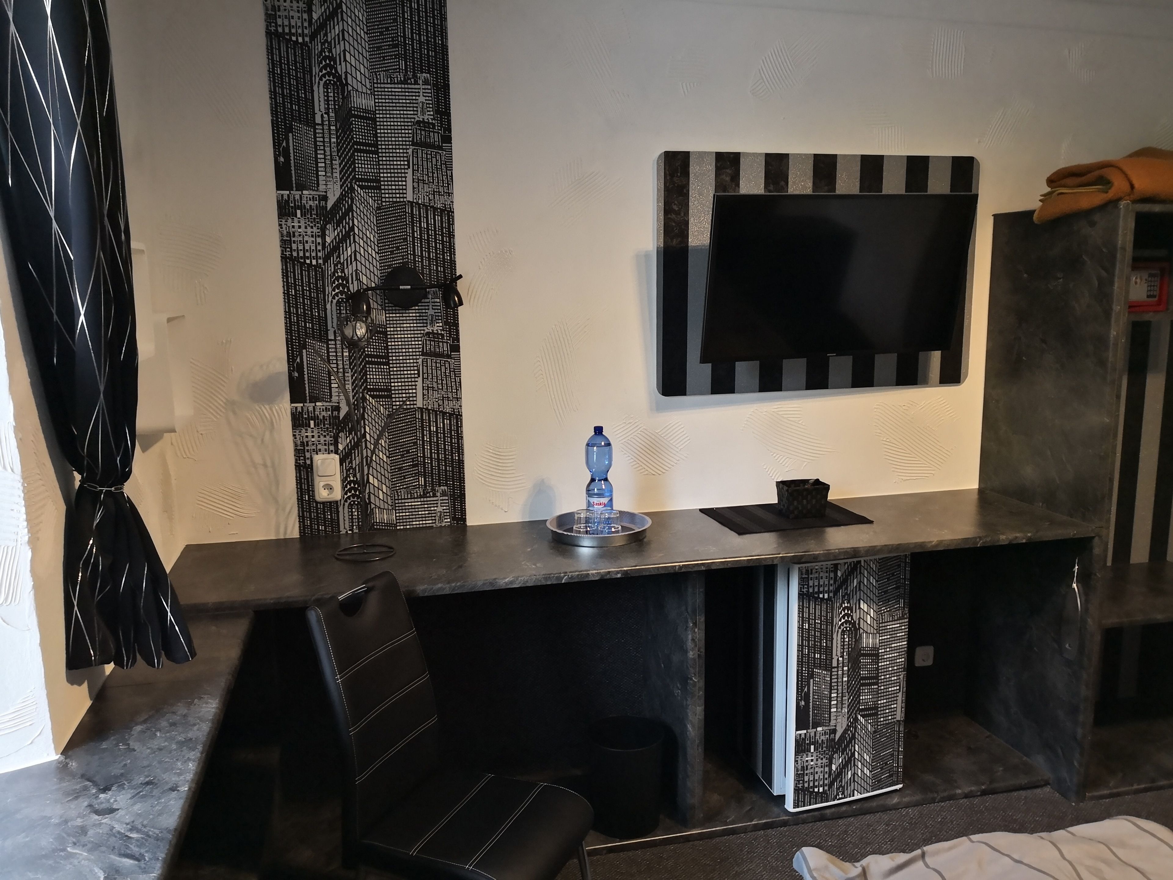 A modern room with a dark wooden desk, a black chair, a flat-screen TV mounted on the wall, and a patterned curtain.