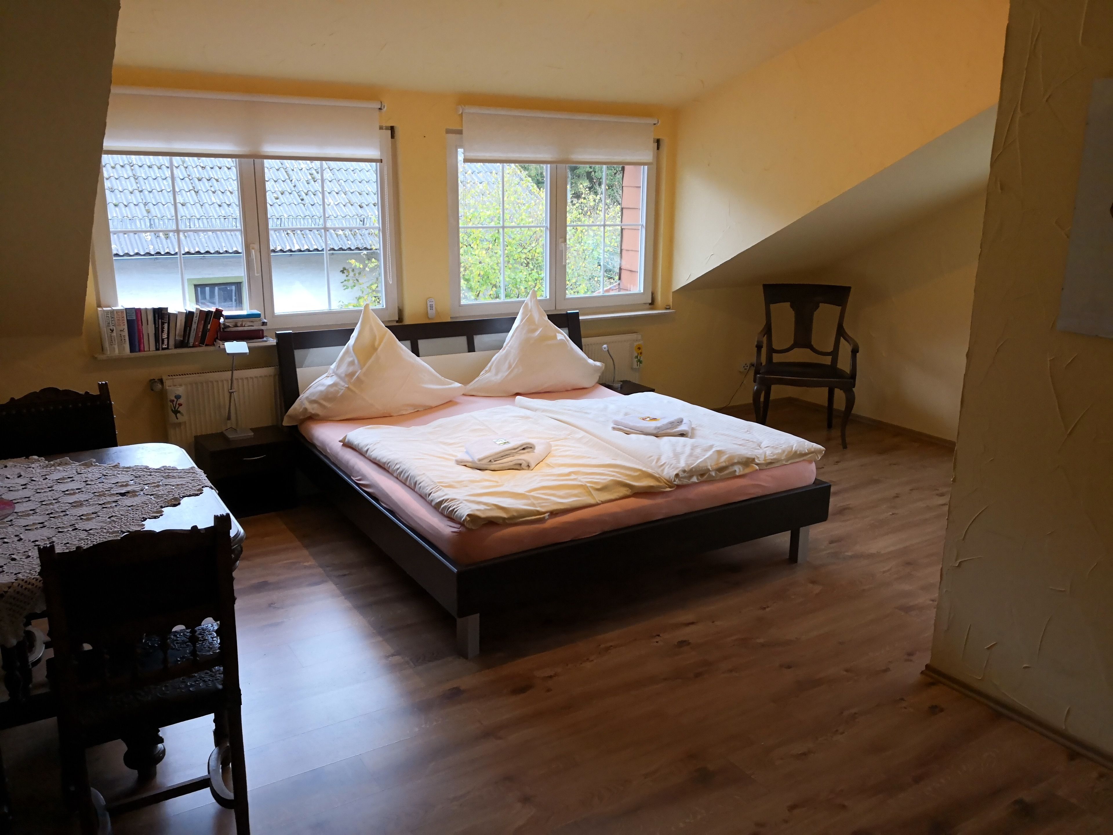 A bedroom with a bed, a desk, and a chair. There are windows with curtains and a radiator on the wall.
