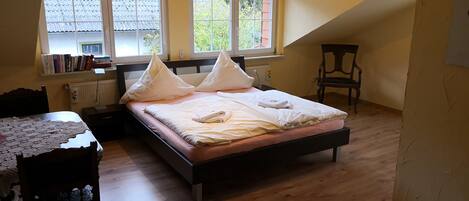 A bedroom with a bed, a desk, and a chair. There are windows with curtains and a radiator on the wall.