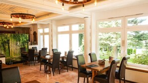 A restaurant with wooden tables and chairs, large windows with a view of greenery, and a rustic ceiling with exposed beams and hanging light fixtures. - Waldgasthaus am Schwarzenbruch (Allenbach)