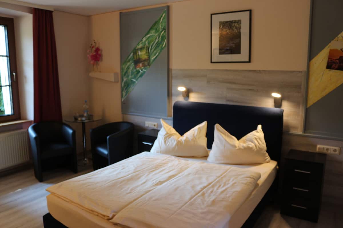 Hotellihuone, jossa on sänky, kaksi tuolia, pieni pöytä, taulu ja lamppu.