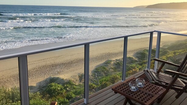 Une terrasse en bois avec une table et des chaises, donnant sur une plage où l’on peut admirer les vagues.